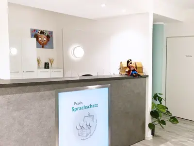Blick in den Eingangsbereich der Praxis auf die Theke mit Sprachschatz-Schild und dem Maskottchen, das auf der Ablagefläche sitzt. Ein türkiser Verlauf kommt von links in das Bild.