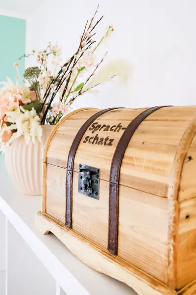 Schatzkiste aus hellem Holz mit Aufschrift "Sprachschatz" steht vor Vase mit beigen und lachsfarbenen Blumen auf einer weißen Ablage.