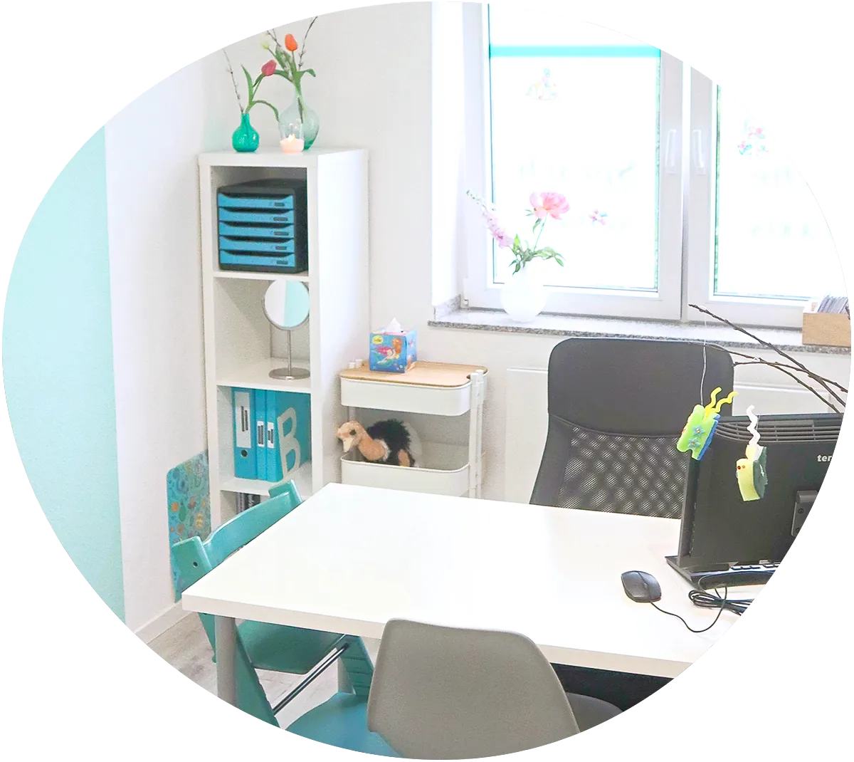 Leicht schräger Blick von oben auf einen Besprechungstisch mit Computer, zwei Stühlen und einem Hochstuhl für ein Kind. Die Sitzgruppe steht vor einem hellen Fenster mit zwei Regalen. Die Wand links ist türlkis.